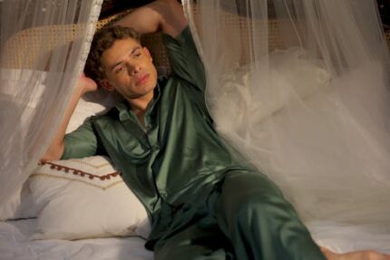 Young man relaxing in bed with canopy, wearing green silk pajamas, looking thoughtful.